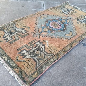 May include: A rectangular area rug featuring a faded orange background with a central blue and pink geometric medallion. The rug displays intricate dark green and cream tribal motifs throughout the field and includes white fringe on both short ends.