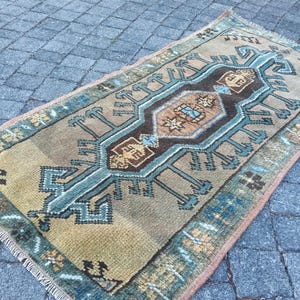 May include: A rectangular area rug featuring a central dark brown diamond medallion with geometric patterns in blue, tan, and cream. The rug has a tan field, a blue border with floral motifs, and short white fringe on the two shorter ends.