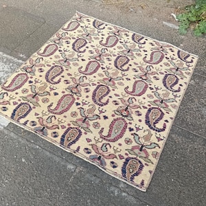 May include: A rectangular rug with a cream-colored background and a repeating paisley pattern in shades of purple, blue, and green. The rug is placed on a gray stone surface, showcasing its intricate design and traditional style.