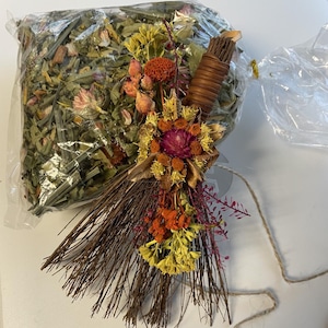 May include: A clear plastic bag filled with dried green herbs and flowers, accompanied by a decorative broom made of thin brown twigs. The broom is adorned with a cluster of dried yellow, orange, and purple flowers tied to its handle.