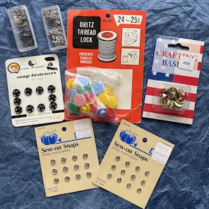 May include: Assortment of sewing supplies including snap fasteners, sew-on snaps, thread lock, and a pack of gold-colored metal eyelets. The packaging is labeled with the brand names "Rich's" and "Dritz".