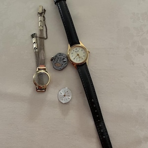 May include: Several vintage watches are displayed, including a gold-toned watch with a black leather band, a gold-toned watch with a mesh band, and watch components. The watch faces are white with black numerals and hands.
