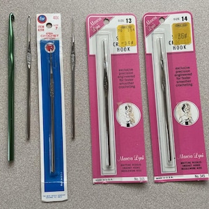 May include: A collection of five crochet hooks, including one green aluminum hook and four silver steel hooks. Two steel hooks are loose, while three are in original packaging labeled Boye size 7, Marcia Lynn size 13, and Marcia Lynn size 14.