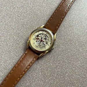 May include: A wristwatch with a brown leather strap and a gold-tone metal case. The watch face features a skeletonized center revealing internal mechanical components, Roman numeral hour markers, and a red second hand with the text Armitron Quartz.