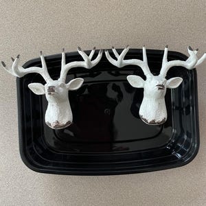 May include: Two white deer head sculptures with large antlers, mounted on a black rectangular tray. The deer heads have a distressed, aged appearance with brown accents on the nose and lower face. The tray is empty.