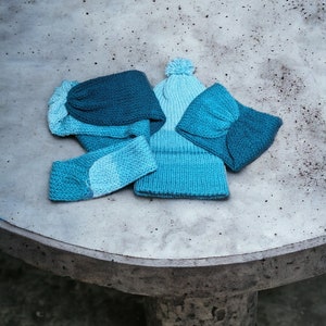 May include: Three knitted hats in shades of blue, with a pom-pom on top of one hat. The hats are arranged on a grey concrete surface.