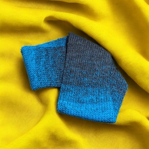 May include: A pair of blue and grey knitted socks with a gradient pattern. The socks are folded and placed on a yellow fabric background.