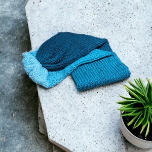 May include: Two knitted hats in shades of blue, one a darker blue and the other a lighter blue, are lying on a grey concrete surface.