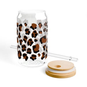 May include: A clear glass can-shaped tumbler featuring a brown and black leopard print pattern. The set includes a matching clear glass straw and a round bamboo lid with a silicone seal and a circular opening for the straw to pass through.