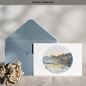 May include: A white greeting card featuring a circular watercolour illustration of a mountain landscape with a lake and trees. The text Happy Father's Day is written in gold cursive script over the image. A blue envelope is positioned behind the card.