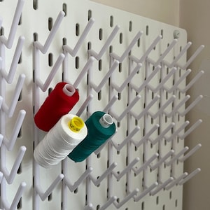 Könnte beinhalten: Eine weiße Lochwand mit weißen Plastikhaken, an denen Spulen mit rotem, weißem und grünem Garn hängen.