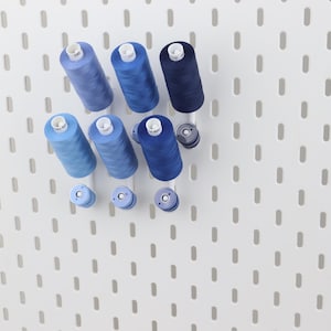 May include: A collection of six cylindrical sewing thread spools in varying shades of blue, ranging from light sky blue to deep navy, arranged on a white pegboard. Three smaller matching blue bobbins are positioned below the larger thread spools.