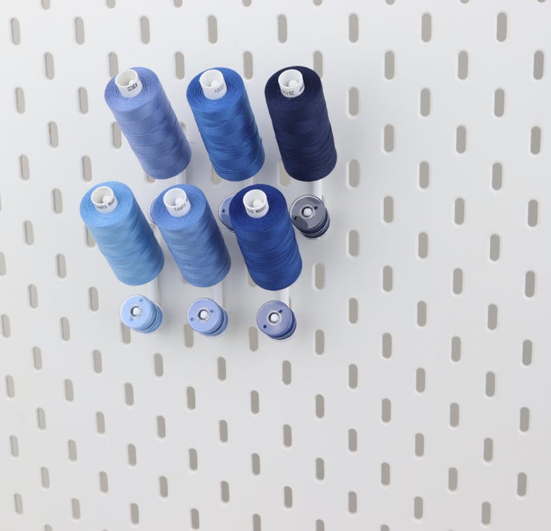 May include: A collection of six cylindrical sewing thread spools in varying shades of blue, ranging from light sky blue to deep navy, arranged on a white pegboard. Three smaller matching blue bobbins are positioned below the larger thread spools.