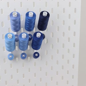 May include: A white pegboard wall organizer holding six spools of sewing thread in varying shades of blue, ranging from light sky blue to deep navy, along with three matching smaller bobbins arranged on white pegs extending from the board.