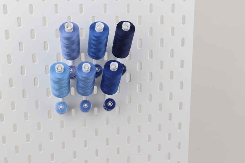 May include: A white pegboard wall organizer holding six spools of sewing thread in varying shades of blue, ranging from light sky blue to deep navy, along with three matching smaller bobbins arranged on white pegs extending from the board.
