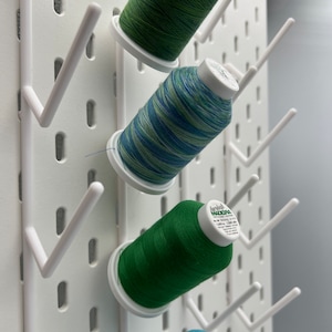 Pu&ograve; includere: Tre bobine di filo sono organizzate su una parete bianca con un pannello forato. Le bobine sono verdi, blu e verdi con strisce bianche. Le bobine sono etichettate "Madeira" e "100% poliestere 120 yd."