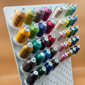 May include: A white plastic storage rack with a variety of colourful spools of thread. The spools are arranged in rows and columns, and they are all different colours, including red, blue, green, yellow, and orange.