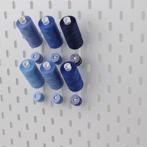 May include: A white pegboard wall organizer featuring three vertical rows of blue sewing thread spools in varying shades of light, medium, and dark blue, with smaller matching thread bobbins positioned directly below each larger spool.