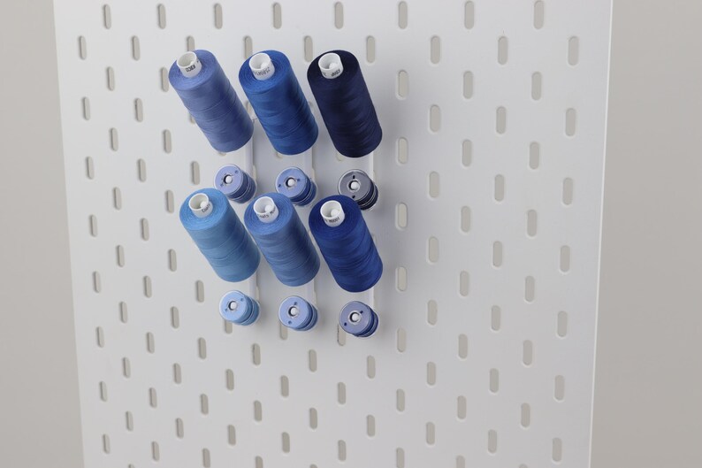 May include: A white pegboard wall organizer featuring three vertical rows of blue sewing thread spools in varying shades of light, medium, and dark blue, with smaller matching thread bobbins positioned directly below each larger spool.