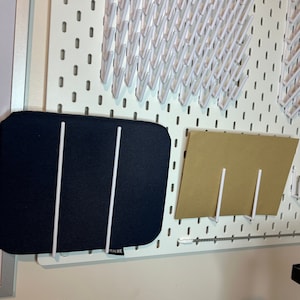 May include: A white pegboard with a black fabric mat and a brown paper mat hanging on it. The fabric mat has two white metal rods attached to it. The paper mat has one white metal rod attached to it. The pegboard has a row of white plastic hooks along the top.