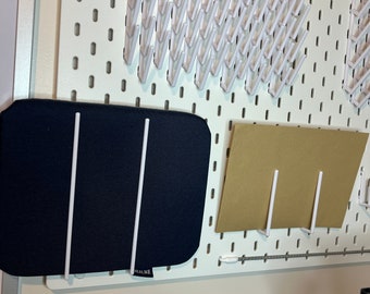 May include: A white pegboard with a black fabric mat and a brown paper mat hanging on it. The fabric mat has two white metal rods attached to it. The paper mat has one white metal rod attached to it. The pegboard has a row of white plastic hooks along the top.