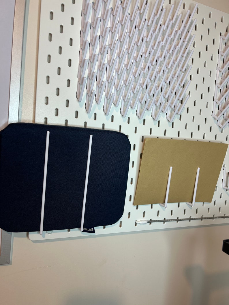 May include: A white pegboard with a black fabric mat and a brown paper mat hanging on it. The fabric mat has two white metal rods attached to it. The paper mat has one white metal rod attached to it. The pegboard has a row of white plastic hooks along the top.