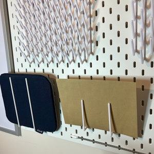 May include: White pegboard with various white hooks and a blue fabric storage pouch with the word "REALIKE" on it. A brown piece of paper is held up by two white hooks.