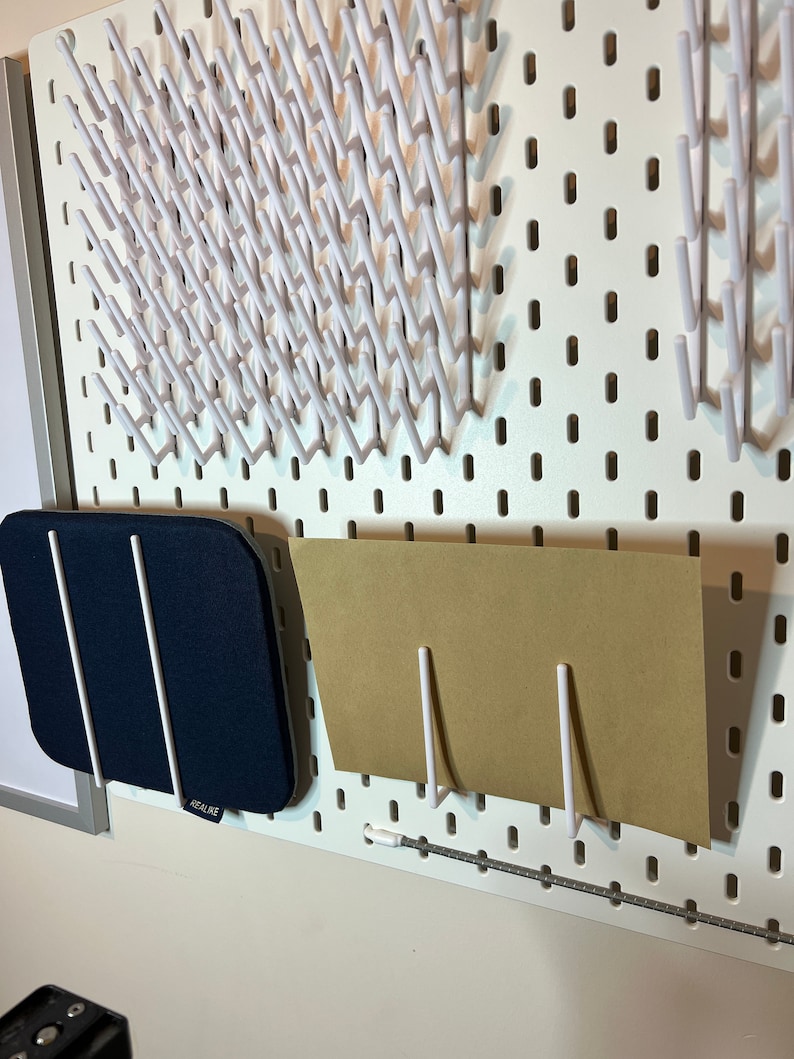 May include: White pegboard with various white hooks and a blue fabric storage pouch with the word "REALIKE" on it. A brown piece of paper is held up by two white hooks.