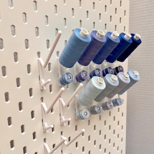May include: A white pegboard with white plastic hooks holding spools of blue thread. The spools are arranged in rows and are different shades of blue.