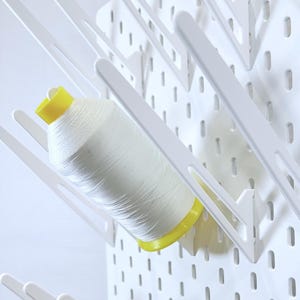 May include: White spool of thread with a yellow base, held in a white pegboard organizer. The pegboard has evenly spaced holes and angled supports for the spools. The background is plain white.