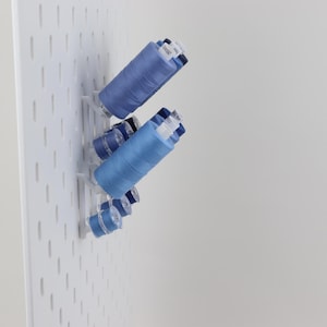 May include: A white pegboard panel features several blue sewing thread spools of varying sizes mounted on white plastic pegs. The spools are arranged in a cluster, displaying different shades of blue thread wound around cylindrical plastic cores.