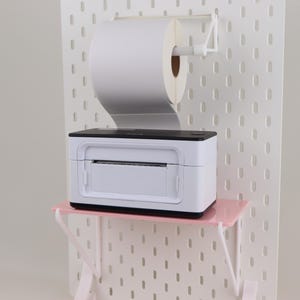 May include: A white and black thermal label printer sits on a small pink shelf attached to a white pegboard. A large roll of white thermal label paper is mounted on a white holder directly above the printer, feeding into the device.