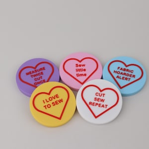 May include: Five circular, flat-topped tokens in purple, pink, light blue, yellow, and white. Each features a red heart outline and red text: MEASURE TWICE CUT ONCE, Sew little time, FABRIC HOARDER ALERT, I LOVE TO SEW, and CUT SEW REPEAT.