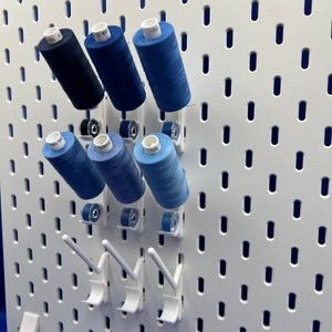 Könnte beinhalten: Eine weiße Lochwand mit sechs Spulen blauen Fadens, die an Haken hängen. Die Spulen sind mit Zahlen beschriftet.