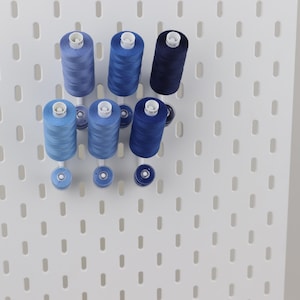 May include: Six spools of sewing thread in varying shades of blue, ranging from light sky blue to deep navy, are mounted on a white pegboard. Each spool is held in place by a white peg, with smaller matching bobbins positioned directly below them.