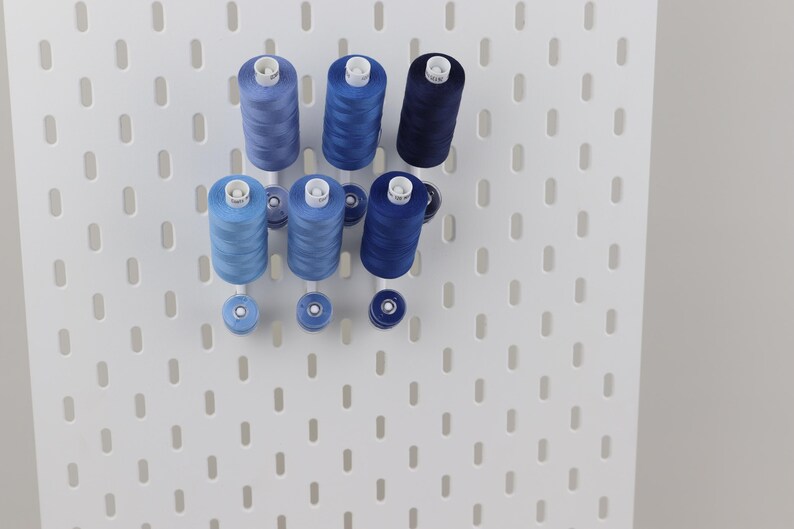 May include: Six spools of sewing thread in varying shades of blue, ranging from light sky blue to deep navy, are mounted on a white pegboard. Each spool is held in place by a white peg, with smaller matching bobbins positioned directly below them.