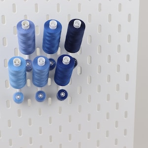 May include: A white pegboard wall organizer holding six spools of sewing thread in varying shades of blue, ranging from light sky blue to deep navy, along with three matching smaller bobbins arranged on white pegs extending from the board.