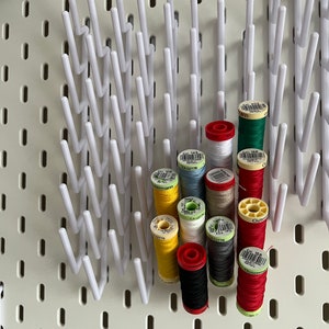 Pu&ograve; includere: Un pannello forato bianco con un porta-bobine bianco che contiene bobine di filo di vari colori, tra cui rosso, verde, giallo, blu, bianco, nero e grigio. Le bobine sono etichettate con il testo "G&uuml;termann" e "CA 02776".