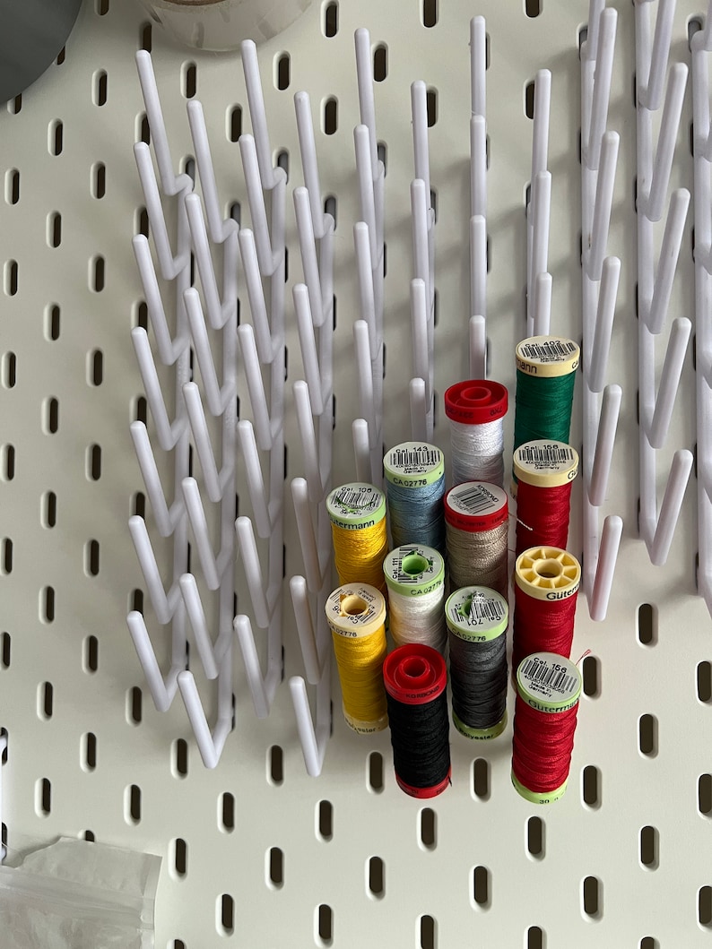 Pu&ograve; includere: Un pannello forato bianco con un porta-bobine bianco che contiene bobine di filo di vari colori, tra cui rosso, verde, giallo, blu, bianco, nero e grigio. Le bobine sono etichettate con il testo "G&uuml;termann" e "CA 02776".