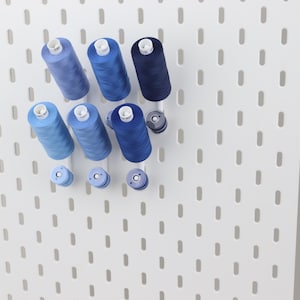 May include: A white pegboard featuring a series of vertical slots, organized with multiple spools of sewing thread in various shades of blue, ranging from light sky blue to deep navy, mounted on pegs to display the thread collection clearly.
