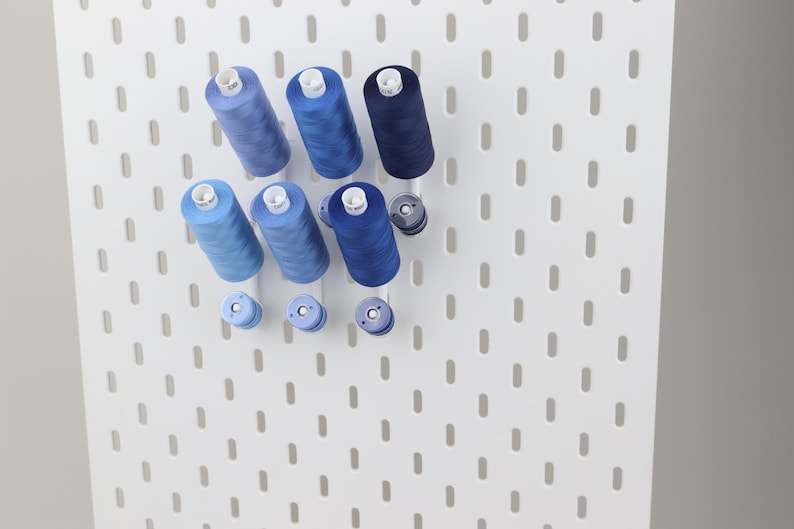 May include: A white pegboard featuring a series of vertical slots, organized with multiple spools of sewing thread in various shades of blue, ranging from light sky blue to deep navy, mounted on pegs to display the thread collection clearly.