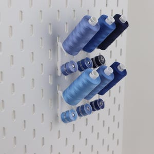 May include: A white pegboard wall organizer featuring a series of blue sewing thread spools in various shades, ranging from light sky blue to deep navy, arranged on horizontal pegs to keep the sewing supplies organized and easily accessible.