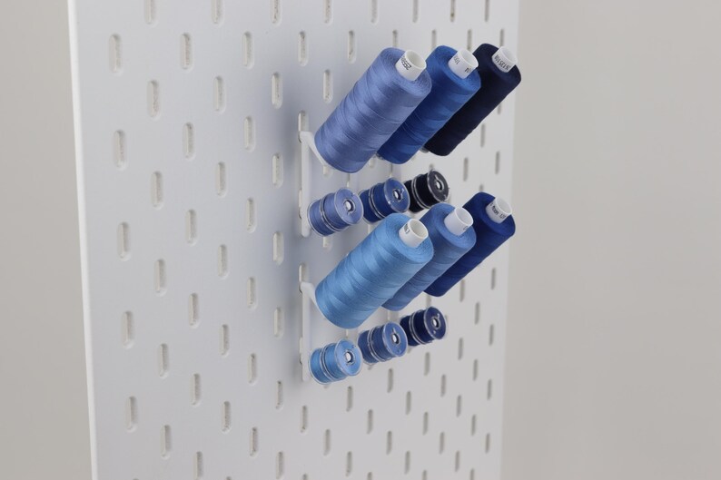 May include: A white pegboard wall organizer featuring a series of blue sewing thread spools in various shades, ranging from light sky blue to deep navy, arranged on horizontal pegs to keep the sewing supplies organized and easily accessible.