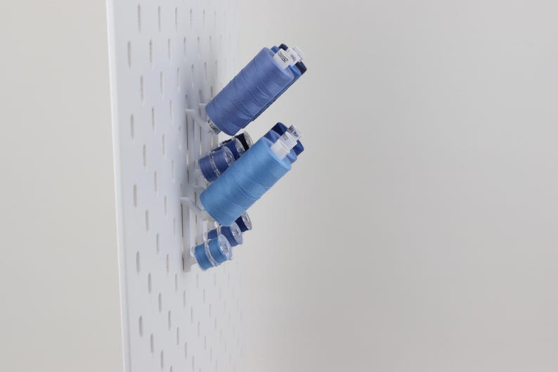 May include: A white pegboard panel features several blue sewing thread spools of varying sizes mounted on white plastic pegs. The spools are arranged in a cluster, displaying different shades of blue thread wound around cylindrical plastic cores.
