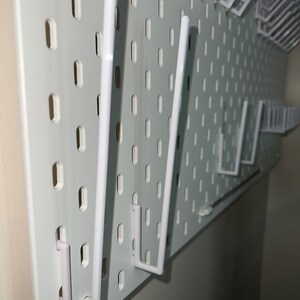 May include: White pegboard with multiple white plastic hooks. The hooks are attached to the pegboard and are in various positions.