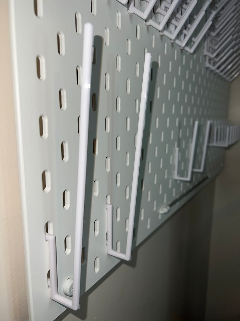May include: White pegboard with multiple white plastic hooks. The hooks are attached to the pegboard and are in various positions.