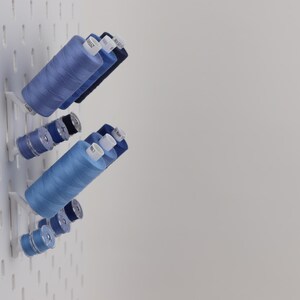 May include: A white pegboard wall organizer holding multiple spools of sewing thread in various shades of blue. The spools are arranged on small white angled pegs, featuring different sizes of thread cylinders with white plastic end caps.
