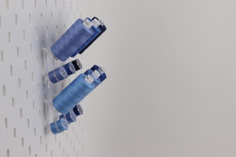 May include: A white pegboard wall organizer holding multiple spools of sewing thread in various shades of blue. The spools are arranged on small white angled pegs, featuring different sizes of thread cylinders with white plastic end caps.