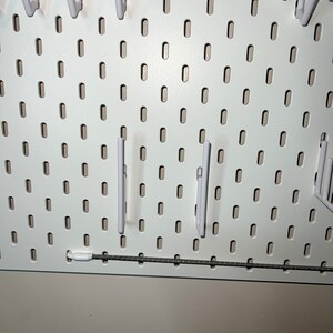 May include: White pegboard with multiple white plastic hooks hanging from it. The pegboard has a silver metal rail at the bottom.