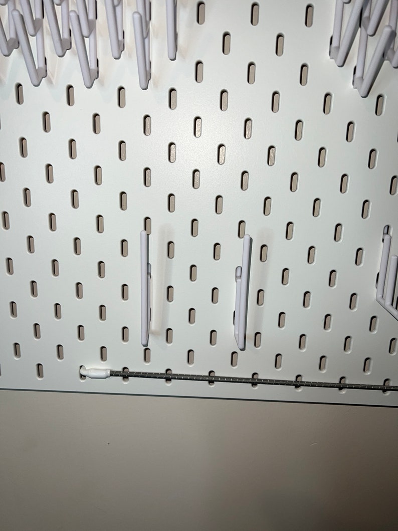 May include: White pegboard with multiple white plastic hooks hanging from it. The pegboard has a silver metal rail at the bottom.